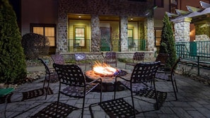 Eine Terrasse im Freien mit einem Feuerkorb, umgeben von Stühlen, ein Steinhaus mit grünen Geländern im Hintergrund und ein gut beleuchteter Eingang.