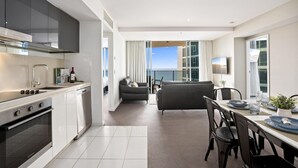 Une cuisine moderne dotée d’un four encastré, un coin repas avec une table dressée pour deux personnes, et une vue sur la mer grâce à de grandes fenêtres.