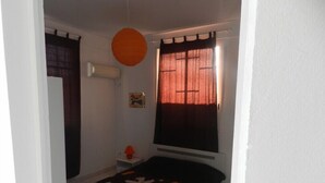 A small, single-bed room with a dark patterned bedspread, a small table with a lamp, and a window with curtains.