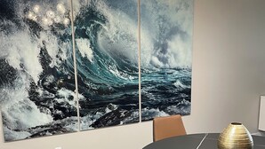 Un coin repas avec une table ronde, des chaises en cuir marron, un vase servant de centre de table et un grand tableau représentant une vague de l’océan accroché au mur.