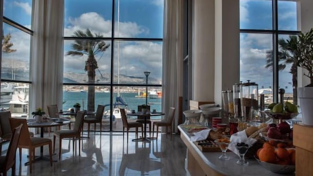 A dining area with a view of the sea, featuring a high table with chairs, a buffet with fruits and pastries, and a large window overlooking the water. - Chios Chandris (Chios)