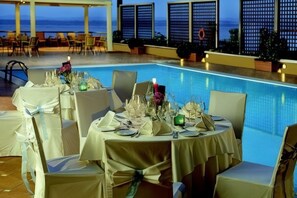 Un área de comedor junto a la piscina con mesas dispuestas para una comida, sillas y vista al océano.
