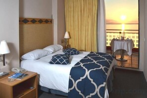Una camera d'albergo con un letto, un comodino, una lampada e vista sul tramonto attraverso una porta finestra.