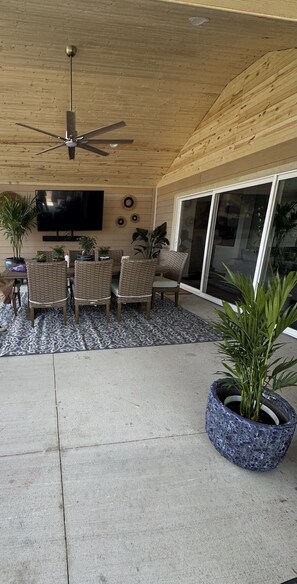 A covered patio area with a ceiling fan, wicker furniture, a flat-screen TV, and potted plants.