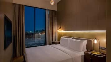 A hotel room with a large bed, a nightstand, a lamp, a view of the city at dusk, and a large window with curtains.