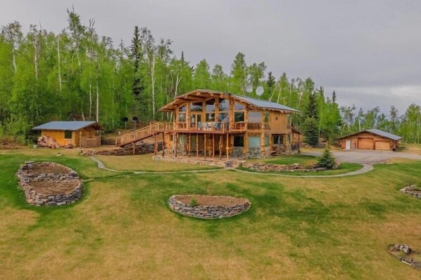 A large log cabin with a deck, surrounded by a well-maintained lawn and stone fire pits.