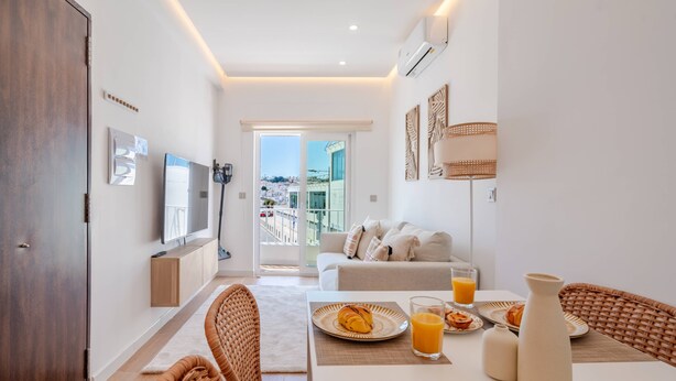 En moderne stue med sofa, spisebord og stole. Der er en balkon med udsigt til bygninger.