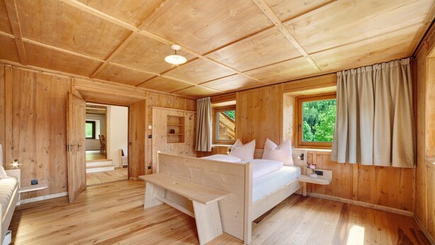 Een houten slaapkamer met een bed, een bankje, een nachtkastje en een raam met gordijnen.