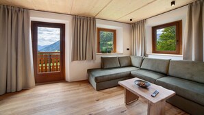 Een kamer met een houten plafond, een groene bank, een houten bijzettafeltje en uitzicht op bergen door een raam.