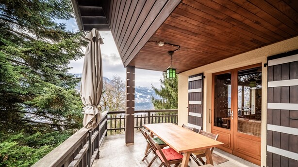 A wooden deck with a table and chairs, a covered area with a view of trees and mountains, and a door with a window. - Chalet 'Franclair' with Mountain View, Shared Garden and Wi-Fi (Samoëns)