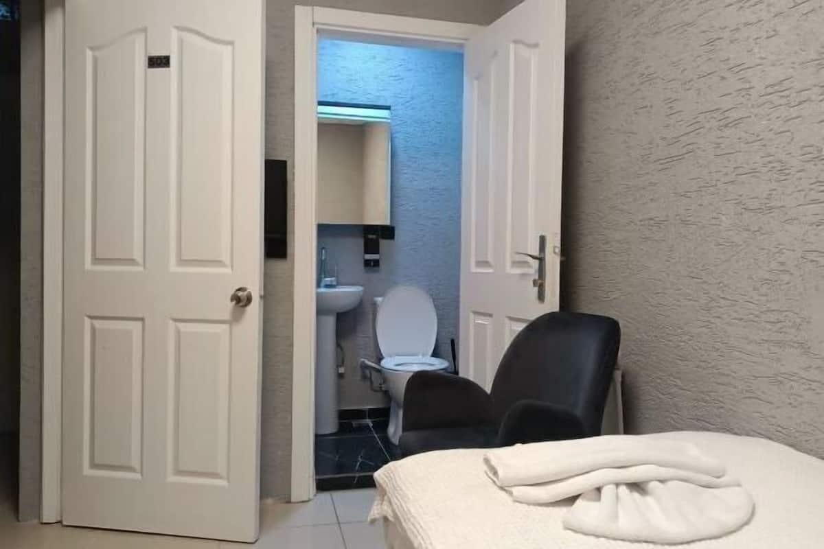 Hotellihuone, jossa on sänky, tuoli, kylpyhuone, jossa on wc ja pesuallas, sekä ovi, joka johtaa toiseen huoneeseen.