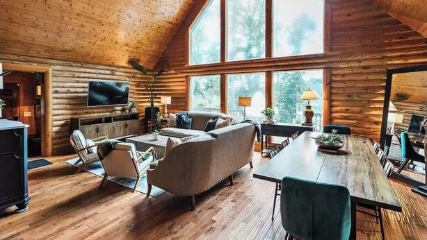 A log cabin interior with a large dining table, a sofa, and a TV mounted on the wall.