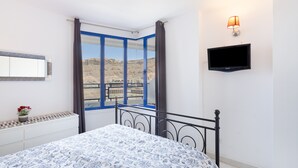 A bedroom with a bed, a TV, a window with curtains, and a view of a mountainous landscape. - Taurito's Sea Sunset View: Sea Views, Shared Pool & Wi-Fi (Taurito)