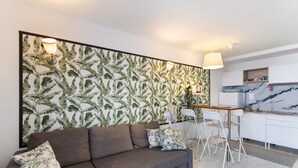 A modern living room with a sofa, a dining area with chairs, a kitchen with white cabinets, and a decorative wall with leaf patterns. - Taurito's Sea Sunset View: Sea Views, Shared Pool & Wi-Fi (Taurito)