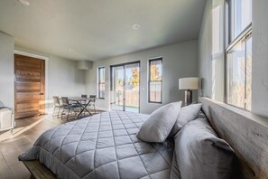 A bedroom with a large bed, a wooden door, a dining area, and large windows. - New 3BR/3BA Condo Steps from Flathead Lake (Lakeside)