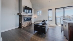 A modern living room with a fireplace, a flat-screen TV, a leather sofa, and large windows. - New 3BR/3BA Condo Steps from Flathead Lake (Lakeside)