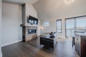 A modern living room with a fireplace, a flat-screen TV, a leather sofa, and large windows. - New 3BR/3BA Condo Steps from Flathead Lake (Lakeside)