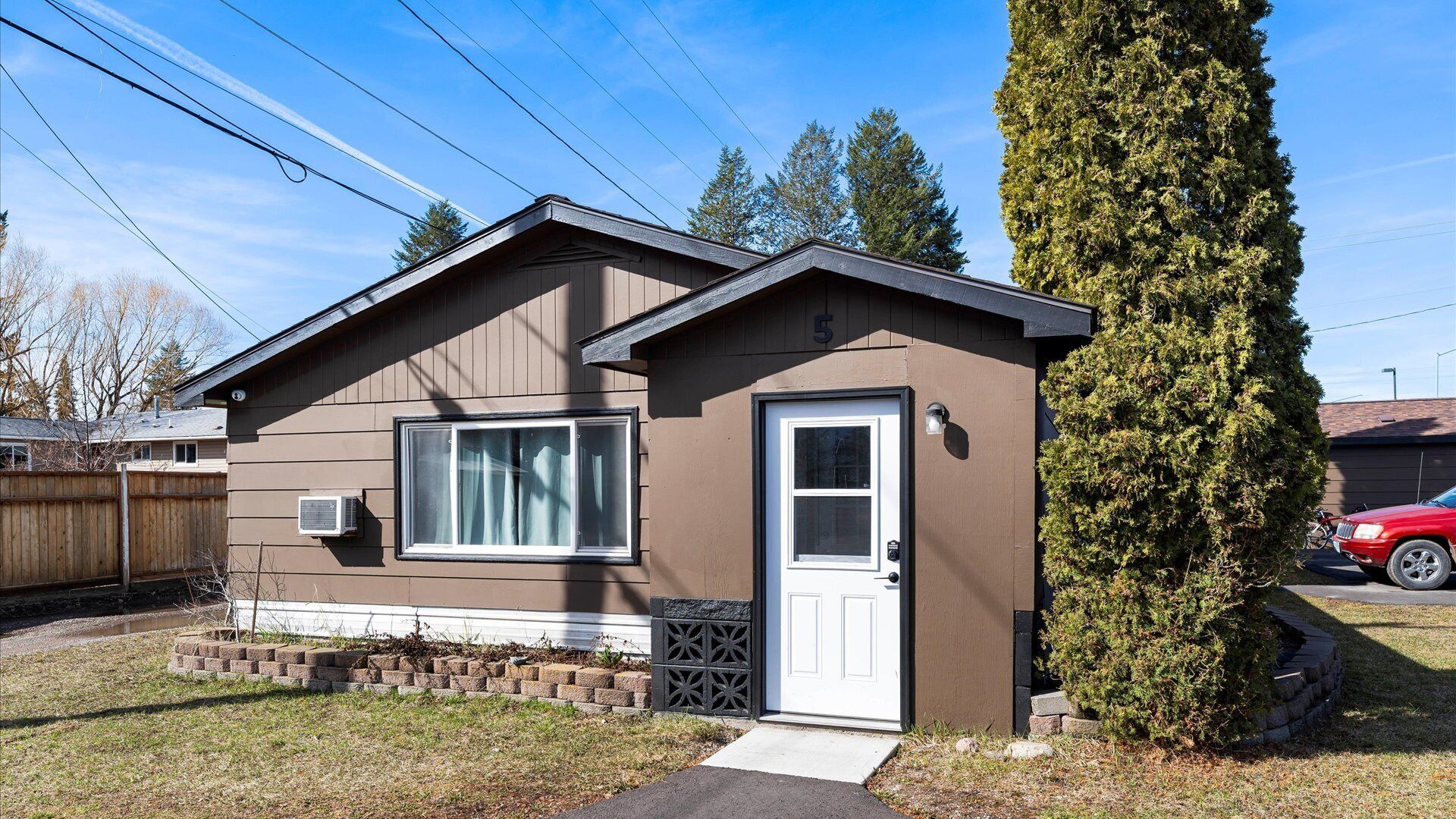 A single-story house with a brown exterior, a white door, and a window with a curtain. There is a driveway leading to the house, and a tall evergreen tree is visible to the right.