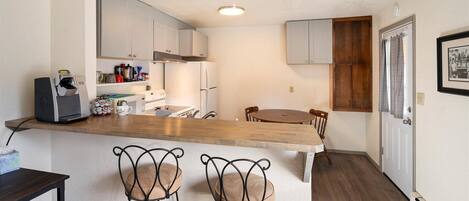 A compact kitchen with white cabinets, a wooden countertop, a coffee machine, and a dining area with a wooden table and metal chairs.