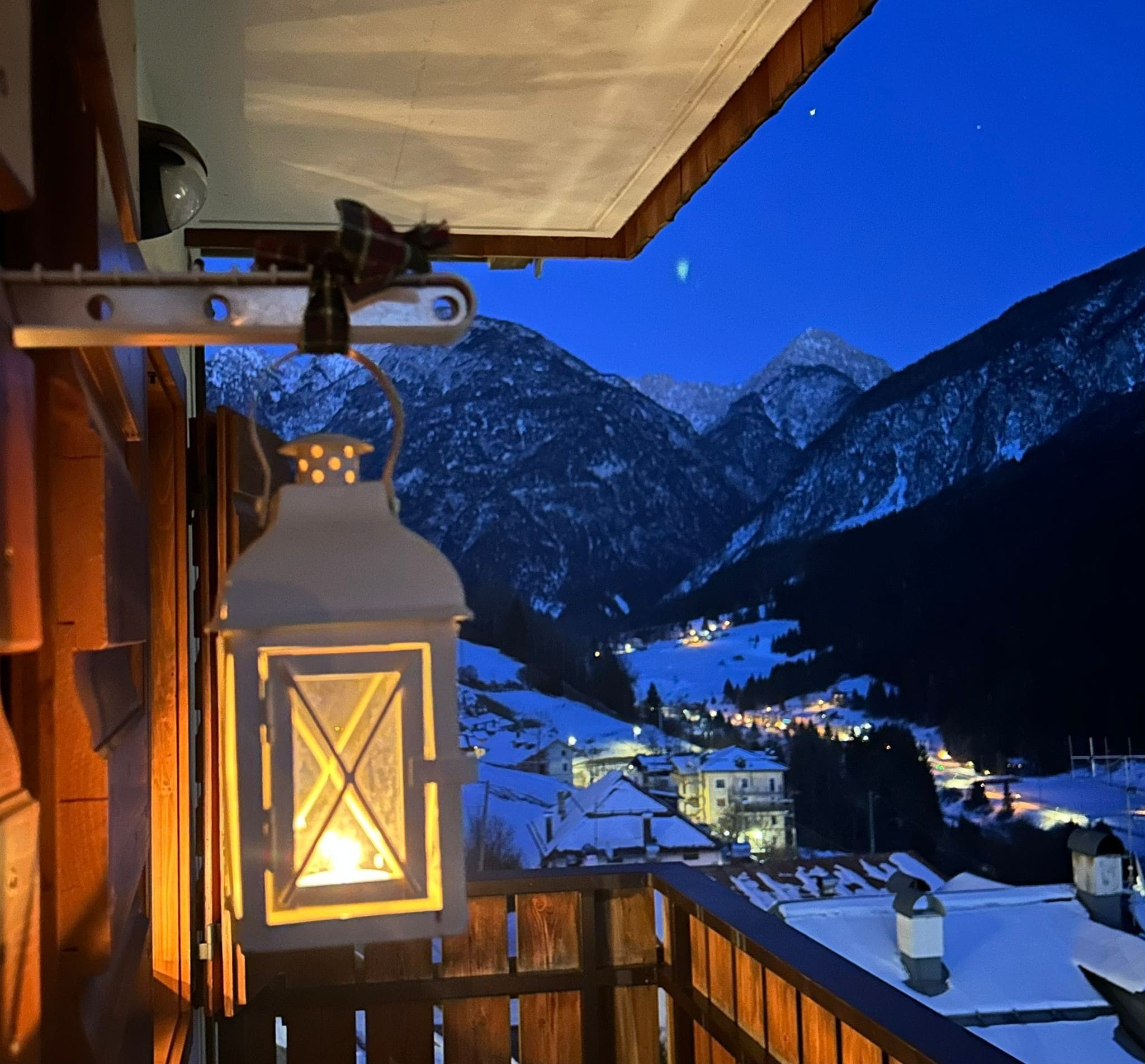 A wooden balcony with a lit lantern, overlooking a snow-covered mountain landscape at night.