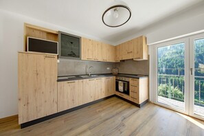 A modern kitchen with wooden cabinets, a built-in oven, and a large window with a view of trees. - Apartment 'Blu' – Mountain Views & Ski Storage (San Pietro di Cadore)