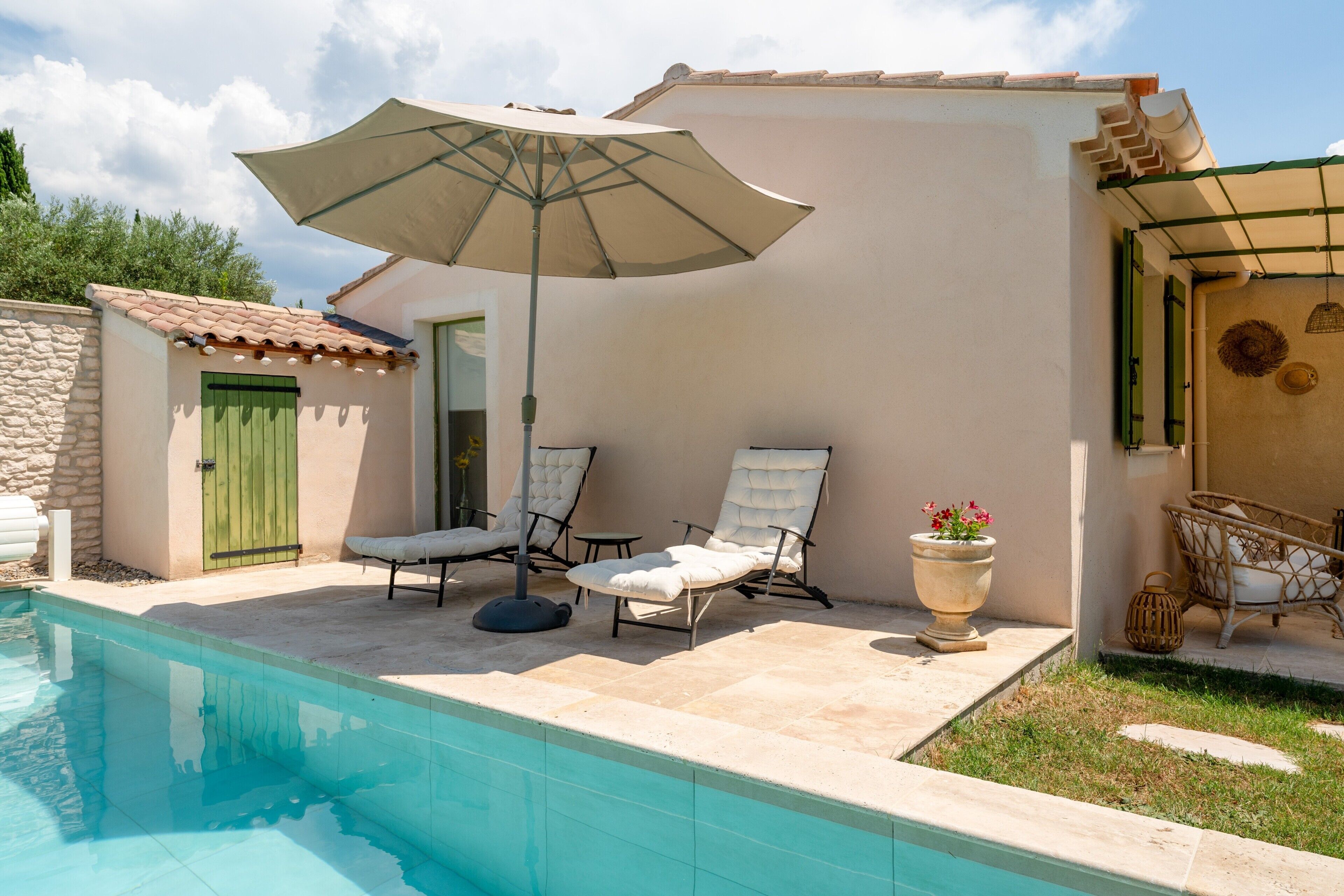 Une piscine avec des chaises longues et un parasol, un mur en pierre et une porte avec un volet vert.
