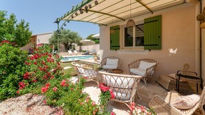A covered outdoor seating area with wicker furniture, a pool in the background, and a building with green shutters. - Lou Cigaloun Holiday Home with Private Pool, Wi-Fi, and Air Conditioning (Bédoin)