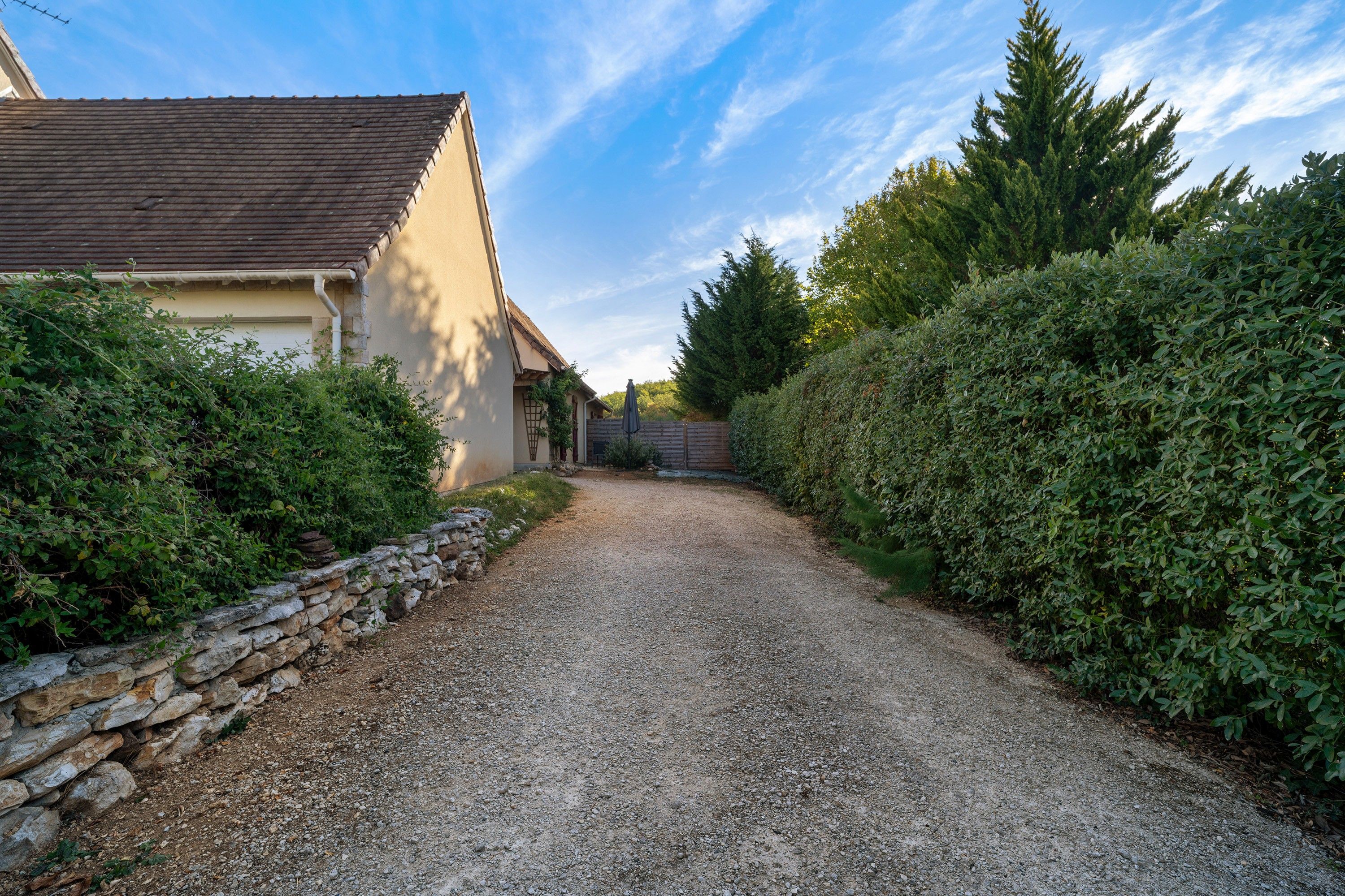 A gravel driveway leading to a house with a stone wall on one side and dense greenery on the other.