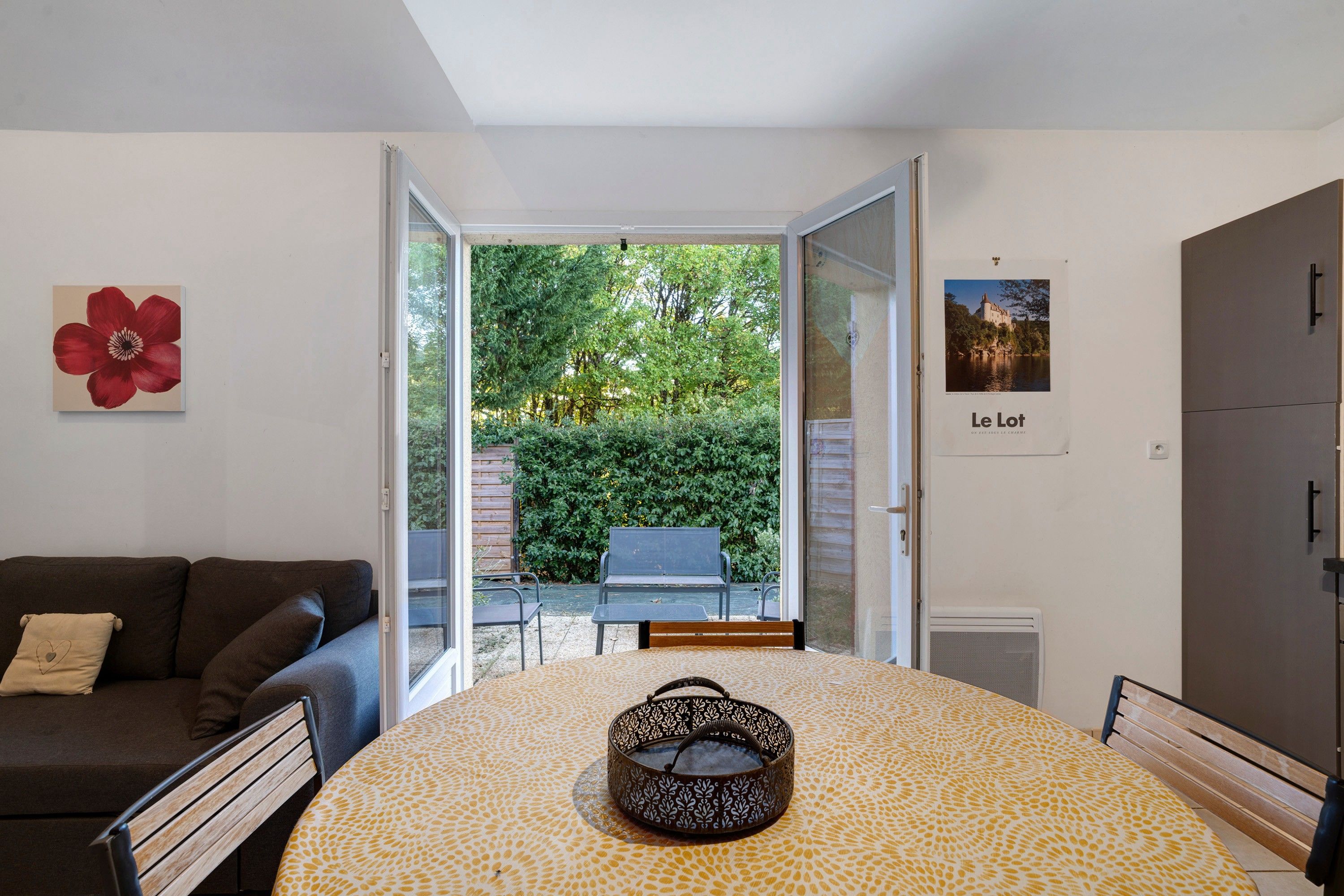 A room with a sofa, a table with a patterned tablecloth, a decorative item on the wall, and a view of a garden through an open door.