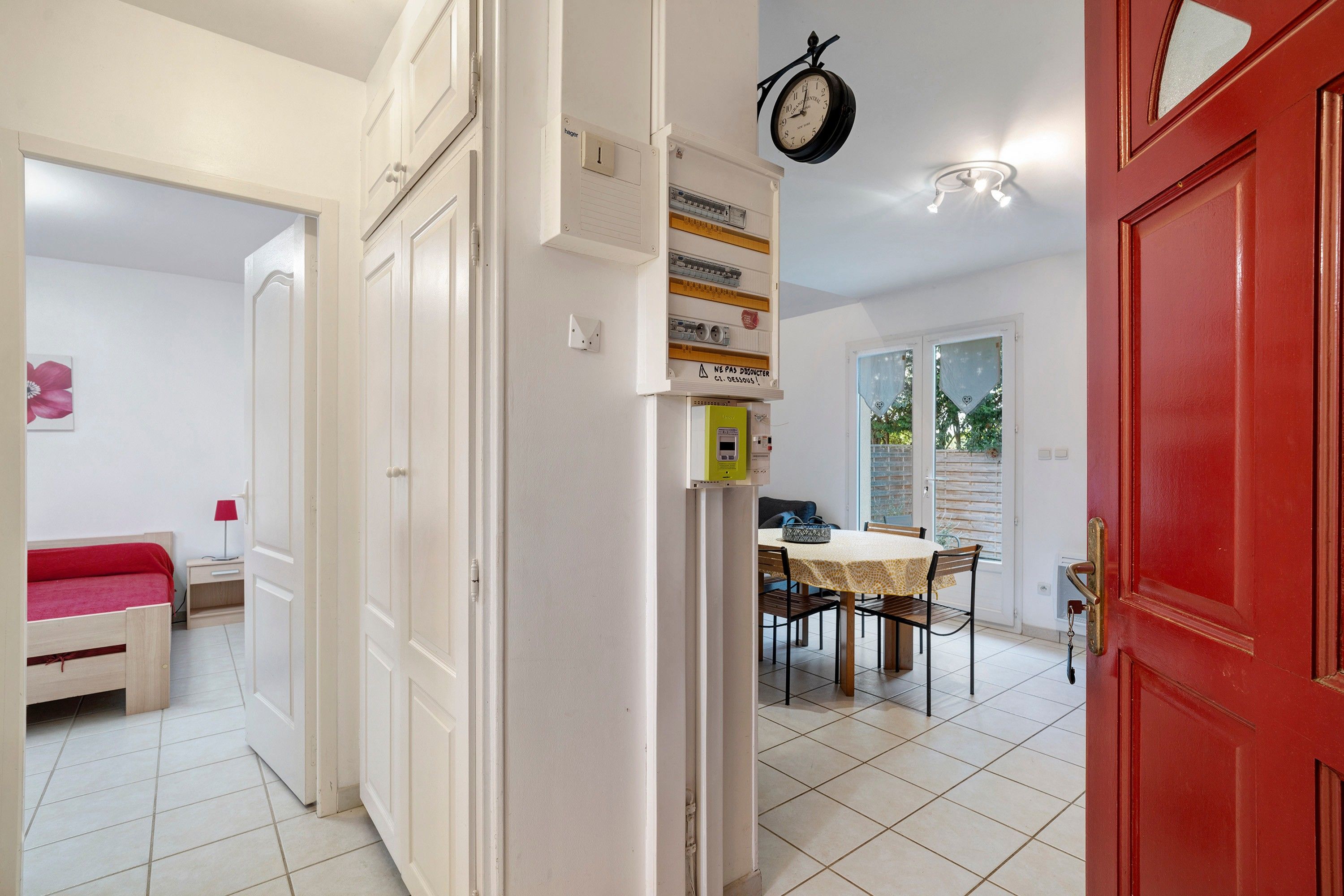 A room with a bed, a dining table with chairs, a red door, and a wall-mounted clock.