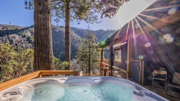 A hot tub with a wooden deck, surrounded by trees and a view of a forested hillside. - River Haus Sequoia | Hot Tub+New+King Beds+Views! - Holiday Home (Springville)