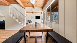 A spacious room with a long wooden table, a staircase, and a large window offering a view of the outdoors. - River Haus Sequoia | Hot Tub+New+King Beds+Views! - Holiday Home (Springville)