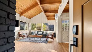 A spacious living room with a wooden ceiling, large windows, a sofa, chairs, and a coffee table. - River Haus Sequoia | Hot Tub+New+King Beds+Views! - Holiday Home (Springville)