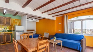 Une cuisine avec des meubles en bois et une table à manger, un coin salon avec un canapé et une fenêtre, et un sol carrelé.