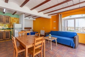 Eine Küche mit Holzausstattung und einem Esstisch, ein Wohnbereich mit Sofa und Fenster, sowie ein gefliester Boden.
