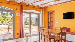 Une pièce comprenant une table à manger en bois, des chaises, un téléviseur fixé au mur et une porte-fenêtre coulissante donnant sur un espace extérieur.