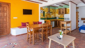 Une pièce comprenant une table à manger en bois, des chaises, une petite table avec une télécommande, un banc avec des coussins et un coin cuisine avec des placards et un pare-feu en carreaux.