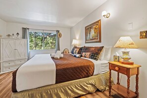 Un dormitorio con una cama grande, una mesita de noche con lámpara, una ventana con cortinas, un armario y un cuadro en la pared.