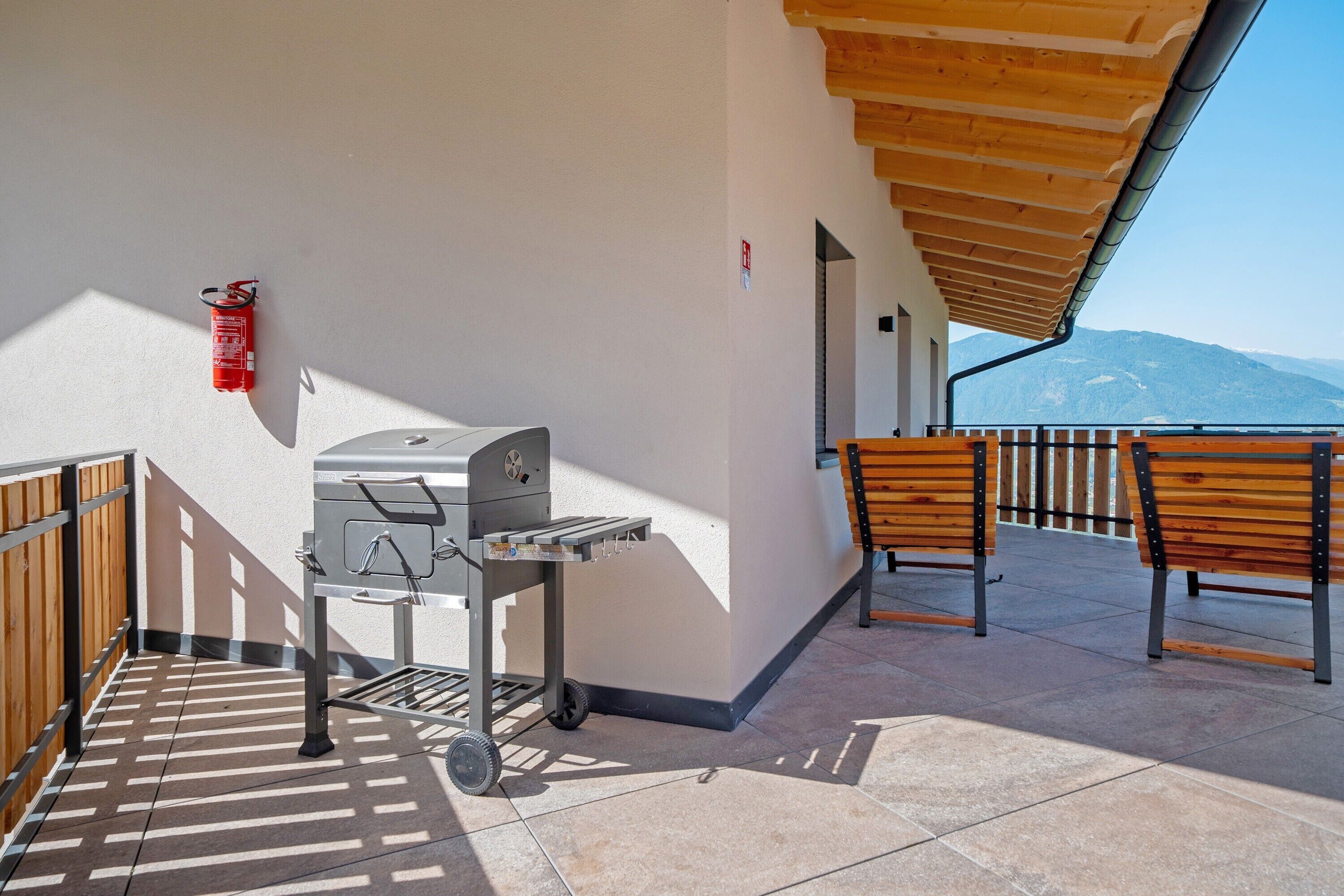 Um terraço com churrasqueira, cadeiras de madeira, extintor de incêndio e vista para montanhas.