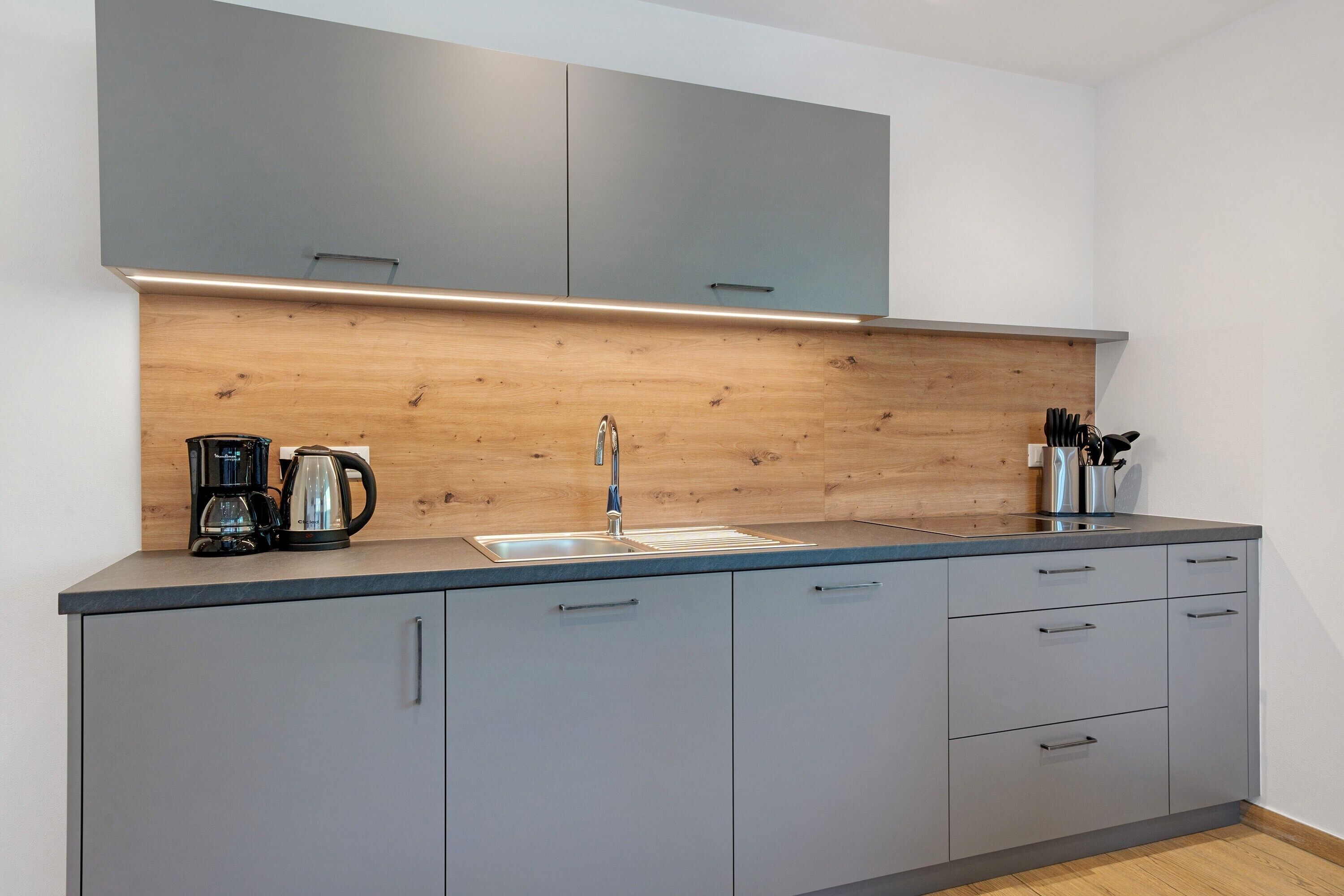 Een moderne keuken met grijze kastjes, een houten spatwand, een roestvrijstalen spoelbak en een koffiezetapparaat.