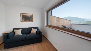 A room with a dark-colored sofa, a small table, a framed picture on the wall, and a large window with a view of mountains.
