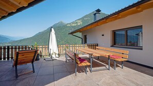A terrace with wooden furniture, a view of mountains, and a building with a chimney.
