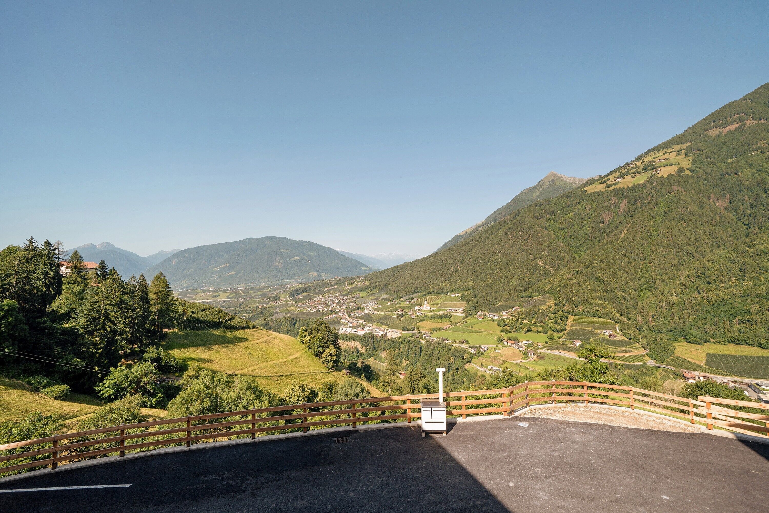 A paved viewing platform with a wooden railing, overlooking a valley with houses and mountains.