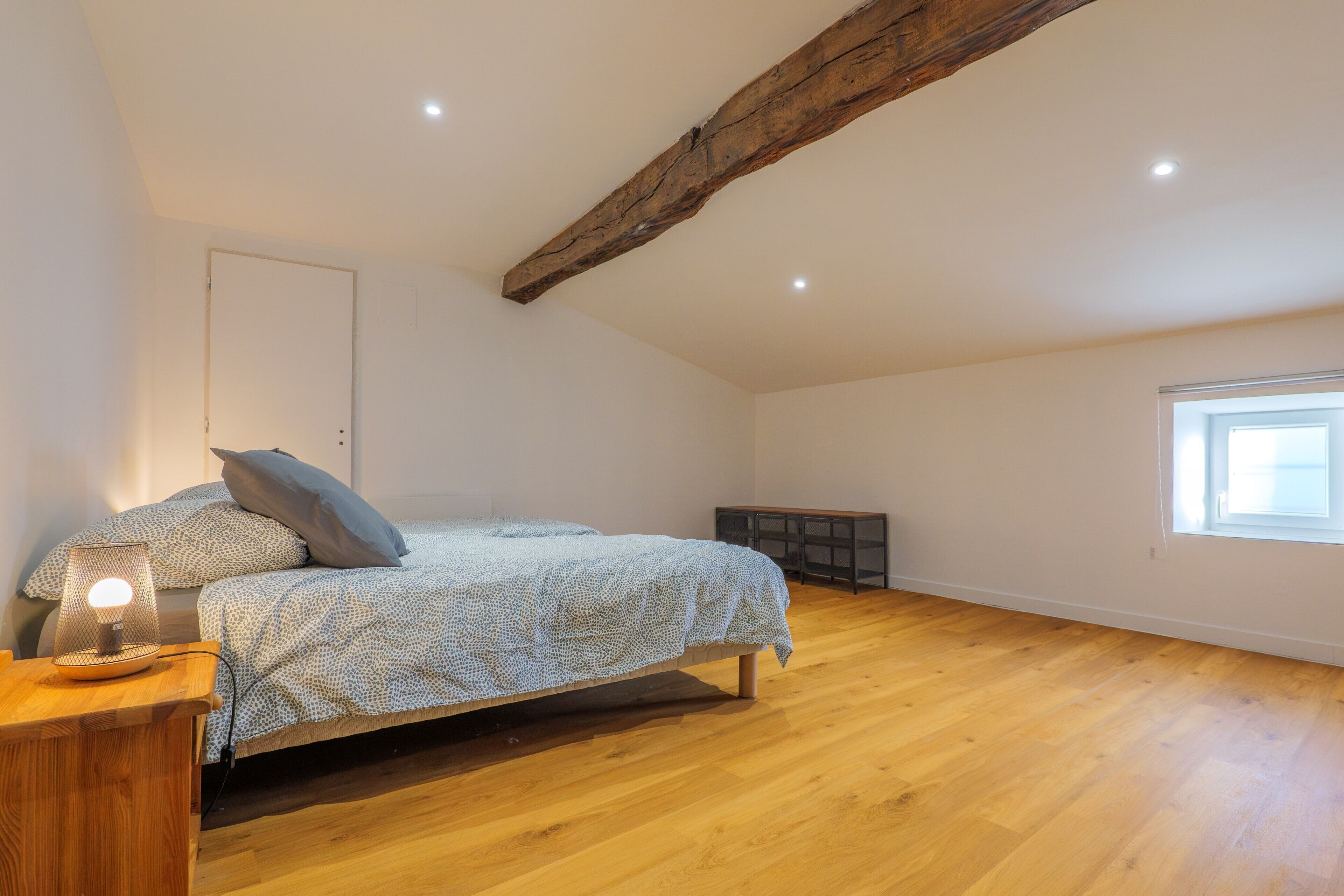Une chambre avec une poutre en bois au plafond, un lit avec une literie à motifs, une table de chevet avec une lampe et une fenêtre.