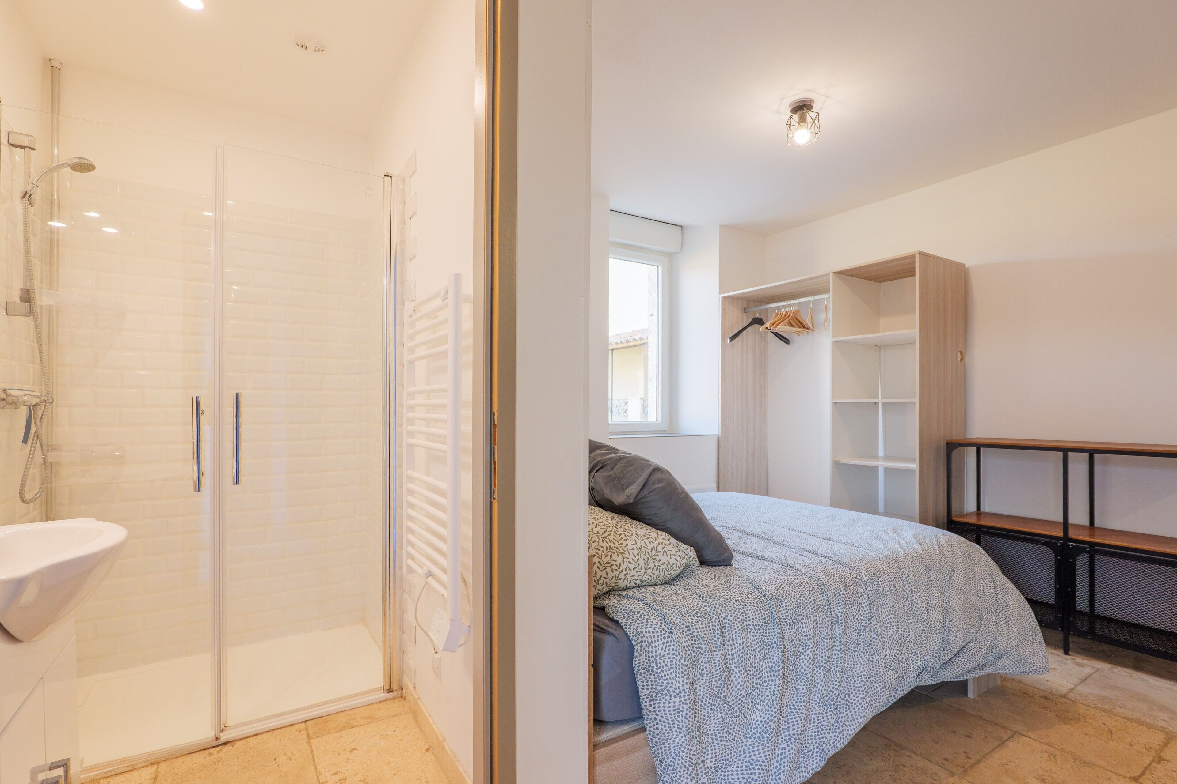A modern bedroom with a glass shower enclosure, a bedside table, a bed with patterned bedding, and a built-in wardrobe.
