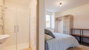 A modern bedroom with a glass shower enclosure, a bedside table, a bed with patterned bedding, and a built-in wardrobe.