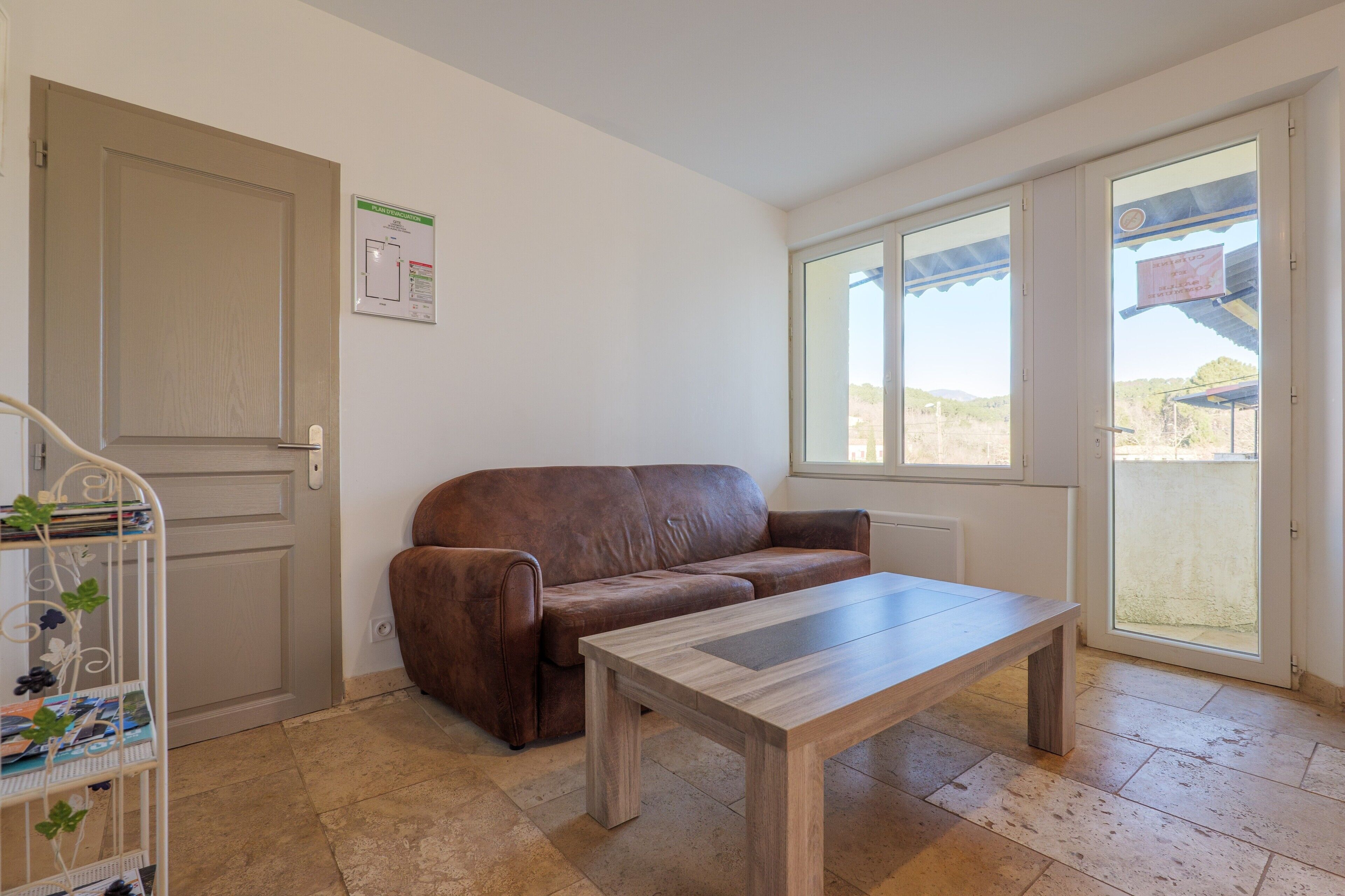 Une pièce avec un canapé en cuir marron, une table basse en bois, une porte blanche, une fenêtre donnant sur un paysage agréable et un sol carrelé.
