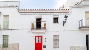 Un bâtiment blanc avec une porte rouge, un balcon orné de plantes en pot et une petite fenêtre munie d’une grille métallique.