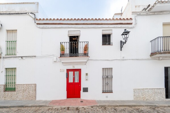 A white building with a red door, a balcony with potted plants, and a small window with a metal grill.