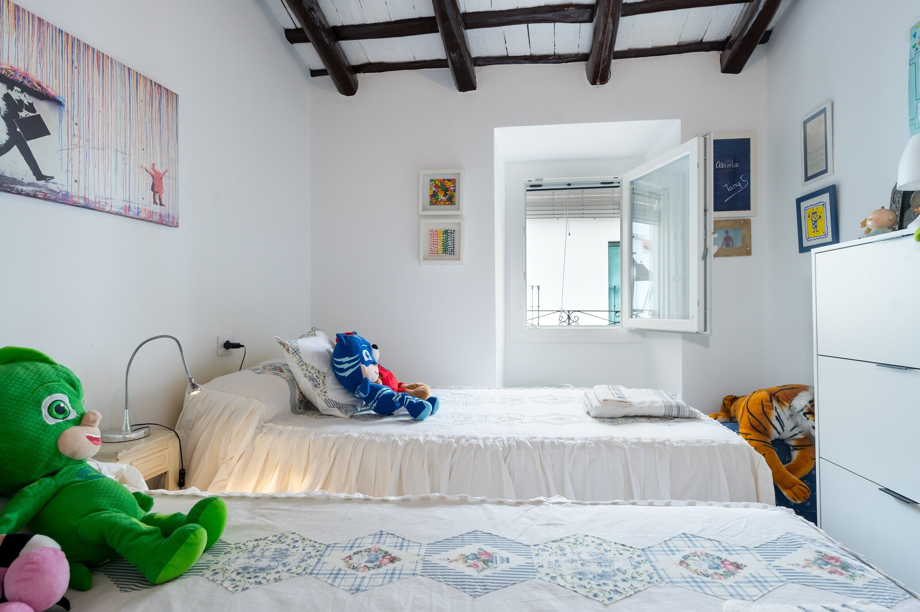 Une chambre à coucher avec un lit, une table de chevet, une lampe, un ours en peluche, un tigre en peluche et un mur décoré d’œuvres d’art.